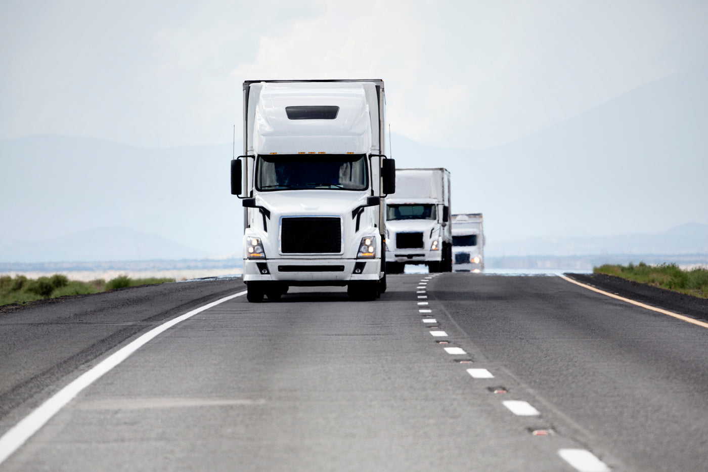 Fleet of white semi-trucks driving on an open highway with mountains in the background.