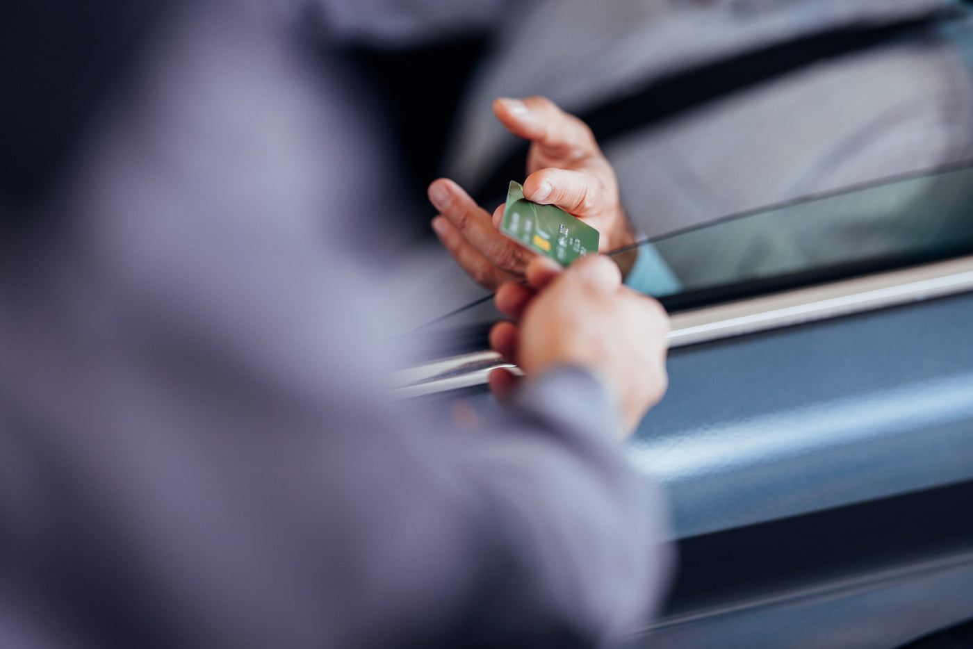 Close-up of a person handing a green credit card through a car window, representing contactless or drive-thru payment.