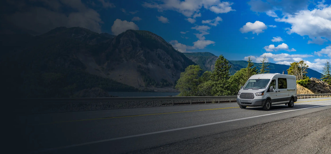 White service van driving along a mountain highway with forested hills and blue sky in the background.