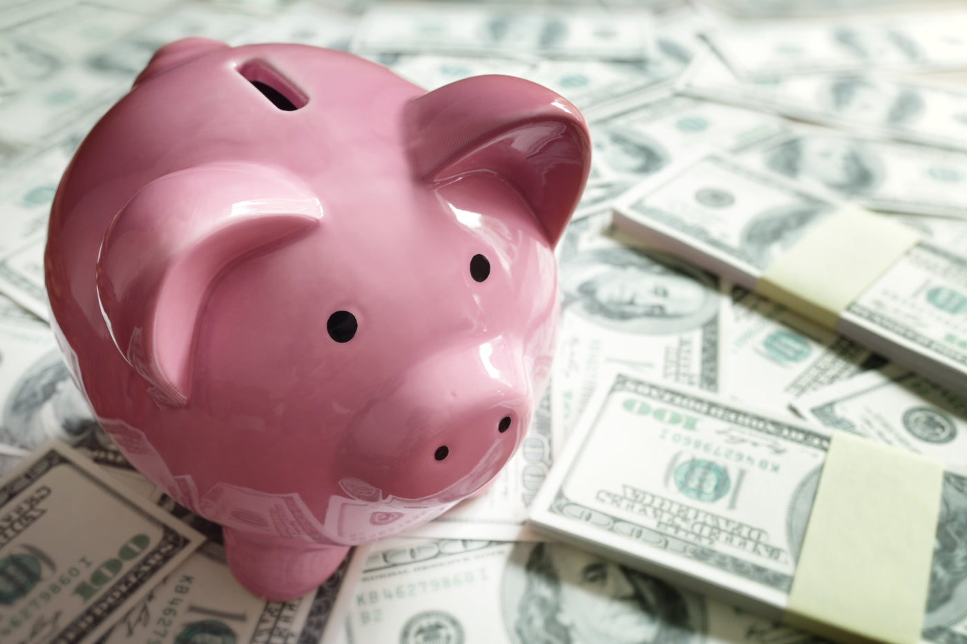 Pink piggy bank sitting on a pile of hundred-dollar bills, symbolizing savings and financial planning.
