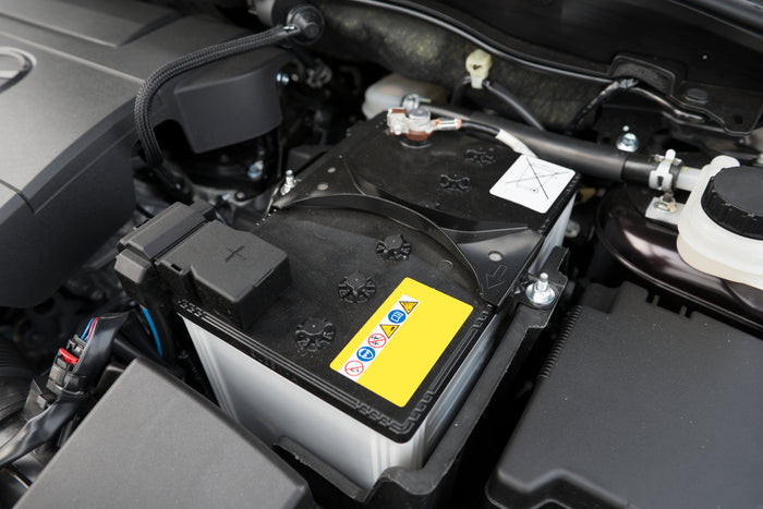Close-up of a car battery under the hood with warning labels and connected terminals visible.