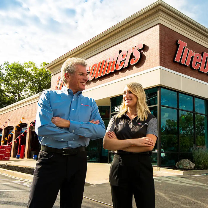 Chip Wood and Anna Wood standing outside a retail store with arms crossed, smiling in front of the building’s branded signage on a sunny day.