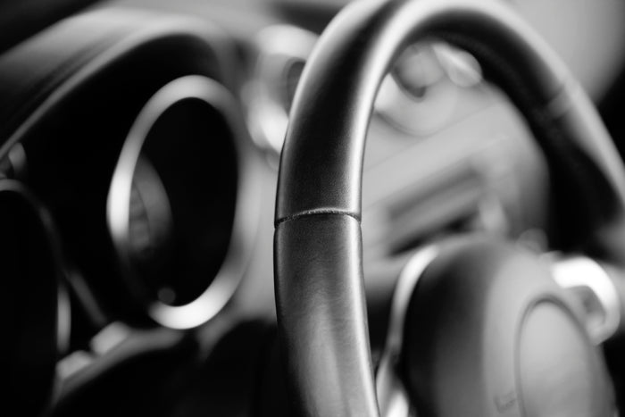 Close-up of a black leather car steering wheel with blurred dashboard in the background.