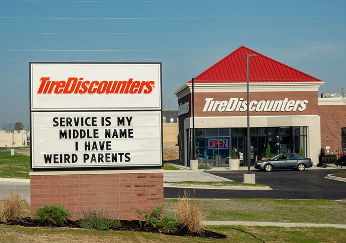 Tire Discounters store with humorous outdoor sign saying “Service is my middle name; I have weird parents” in front of building.