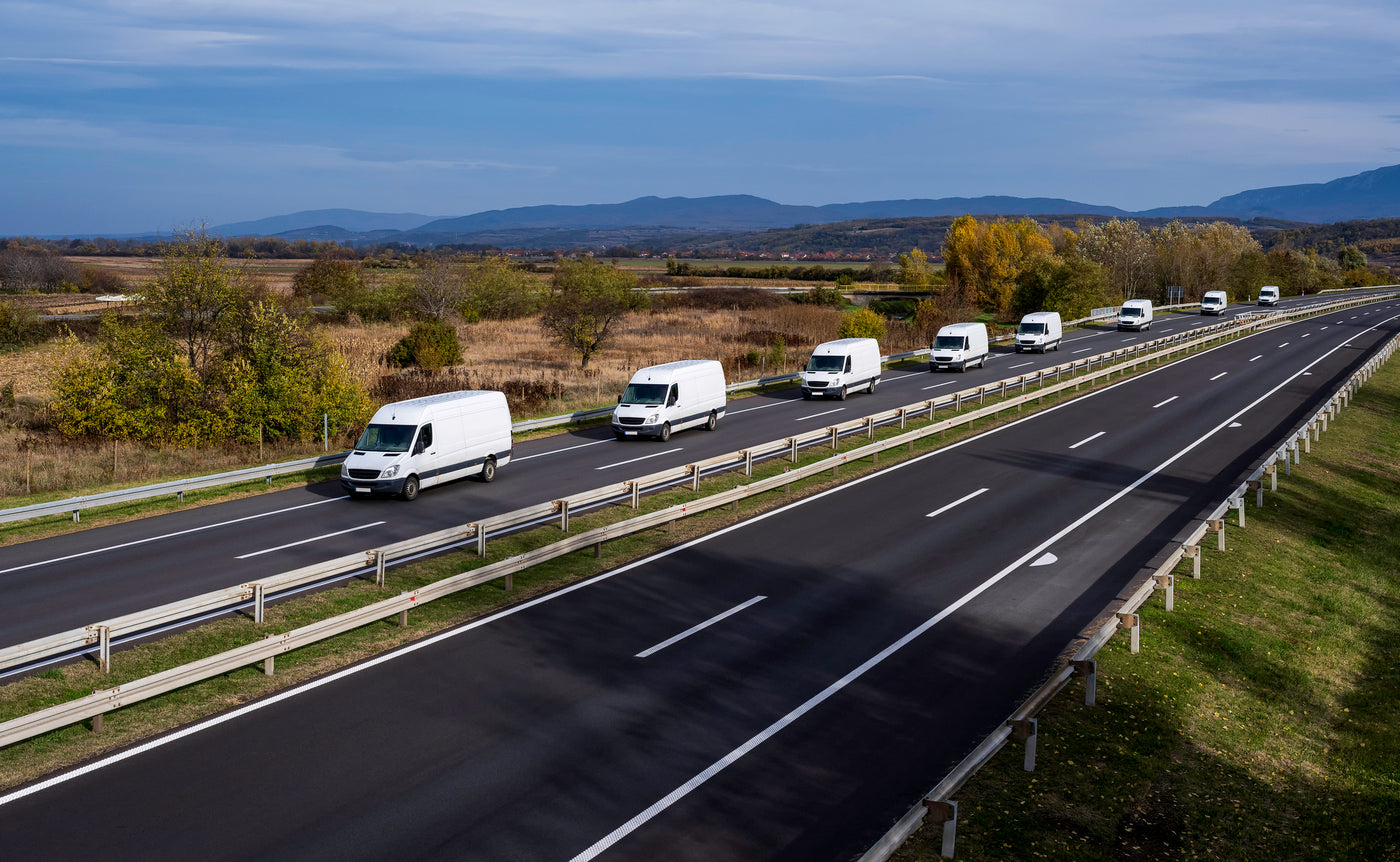 Fleet of white commercial vans driving on a highway through a scenic rural landscape with trees, fields, and distant mountains.