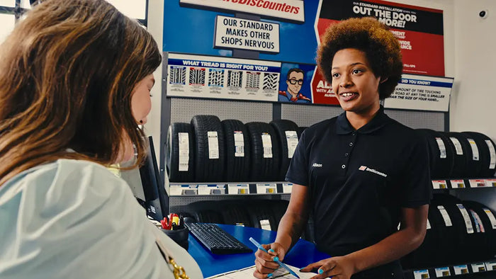 A female Tire Discounters employee in a black polo shirt stands behind a counter, smiling and holding a blue clipboard and pen, while a customer in a light blue top sits across from her. Shelves of tires and store signage are visible in the background.