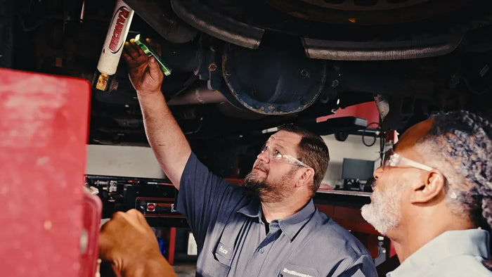 A mechanic in a garage wearing safety glasses and a dark blue work shirt applies a bottle of oil under a car. Another person is partially visible on the right, and a red tool cabinet is on the left.