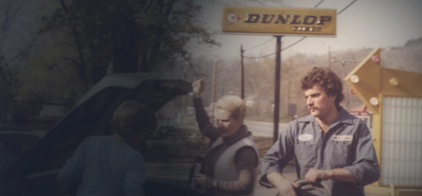 Vintage photo of a mechanic in a blue uniform near a Dunlop Tires sign, assisting two people with a car trunk open roadside.