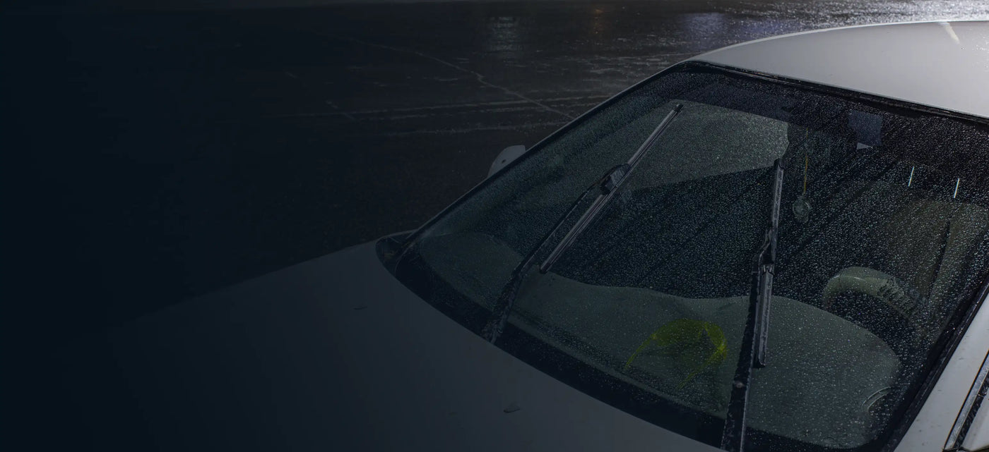 White sedan in a rainy parking lot covered in water droplets with windshield wiper blades in motion.