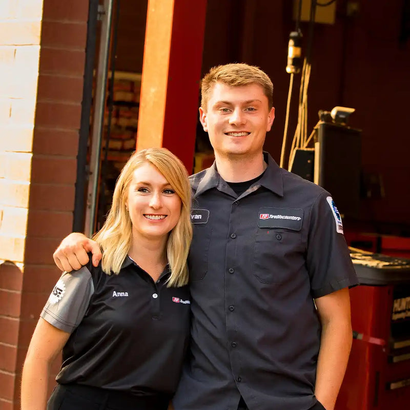 Anna and Evan Wood smiling outside a service bay, wearing branded uniforms and standing arm in arm, showcasing friendly customer service.