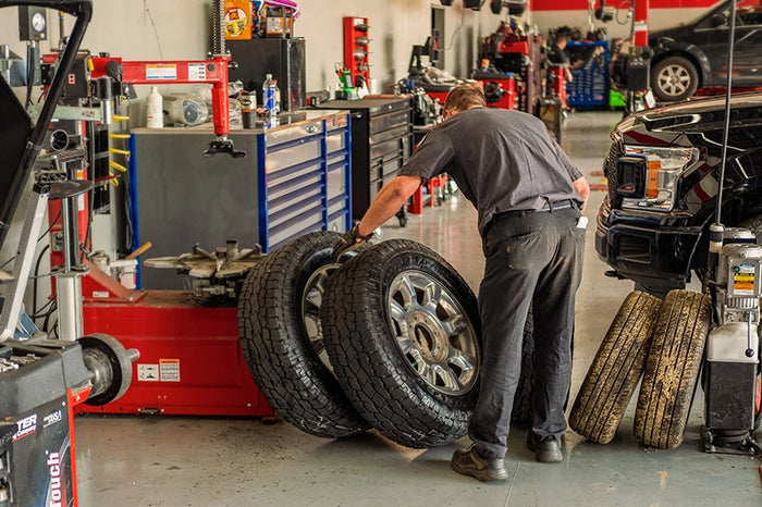 Auto technician handling tires inside a full-service tire shop, surrounded by tools and vehicles in an active garage setting.