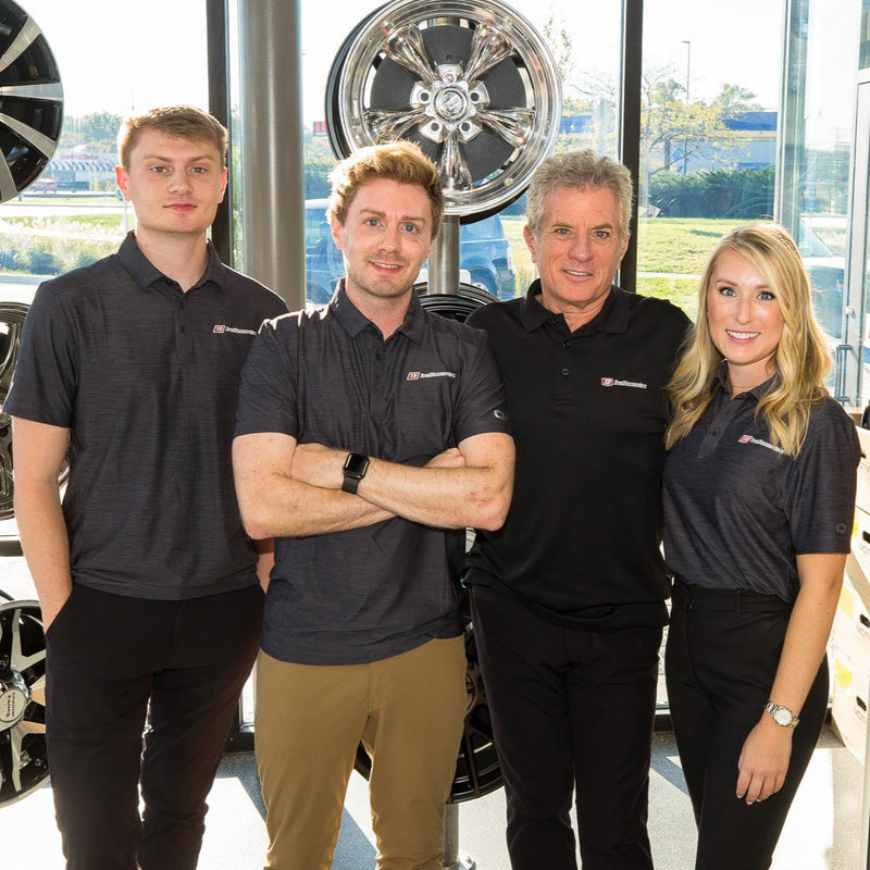 Evan, Steven, Chip, and Anna Wood standing in front of a display of aftermarket wheels in a sunlit showroom, wearing matching branded polos and smiling confidently.