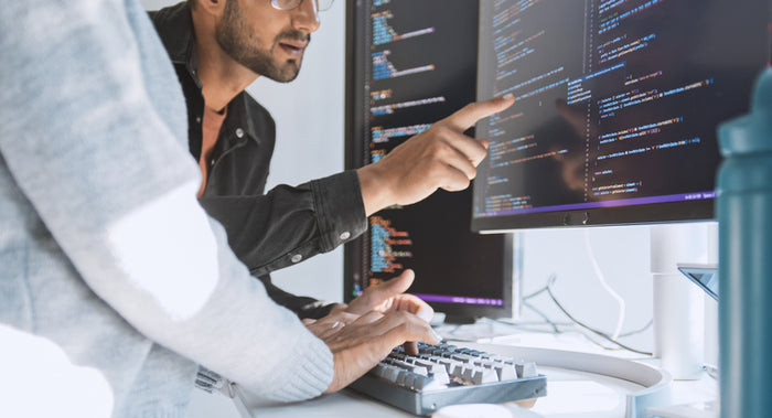Two developers collaborating at a workstation, reviewing and debugging code on dual monitors in a modern software engineering setup.