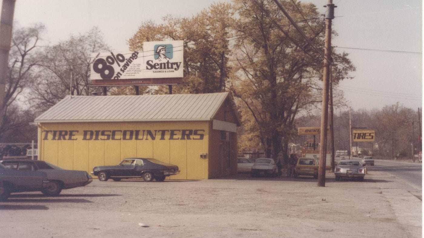 Vintage photo of the original Tire Discounters building with classic cars parked outside and signage for Dunlop Tires and Sentry Savings.