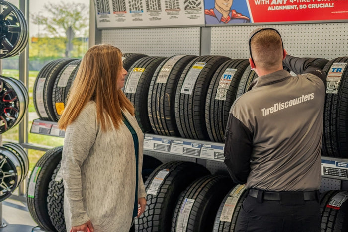 Tire Discounters associate assisting a customer with tire selection in-store, surrounded by labeled tire options and display wheels.