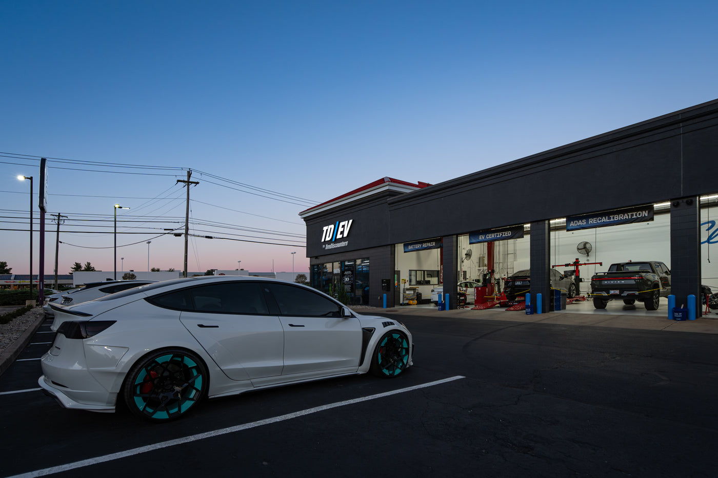 TD/EV by Tire Discounters service center at dusk with a white Tesla parked outside, offering EV repair and ADAS recalibration.