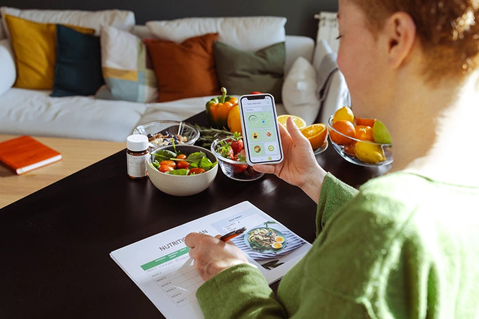 Person tracking nutrition on smartphone while meal planning with fresh fruits and vegetables at home kitchen table.