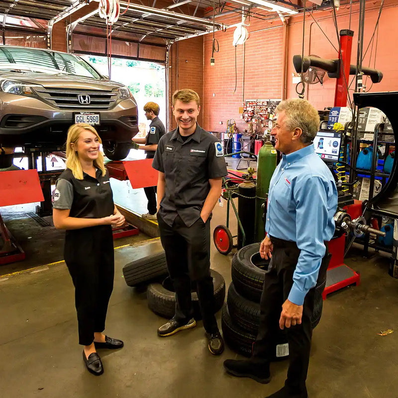 Anna, Evan, and Chip Wood  talking in an auto service bay with a vehicle lifted on a red hydraulic lift in the background, surrounded by tires and shop equipment.